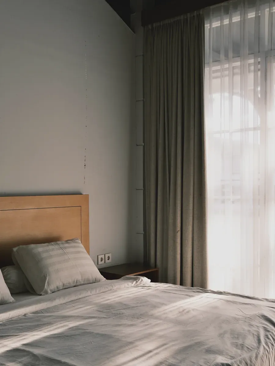 Sunlit bedroom at Tulus Coliving with a wooden headboard, white bedding, and sheer curtains